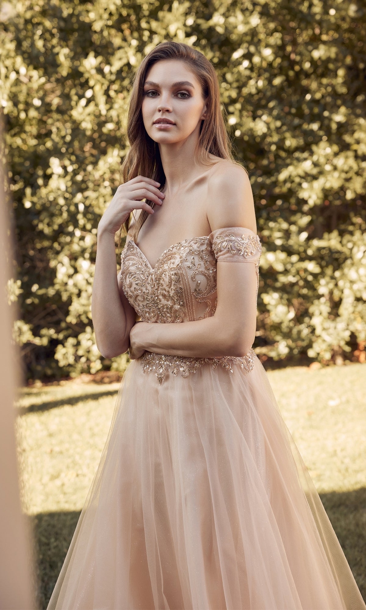 A woman in the Juliet 287 Long Sweetheart Prom Ball Gown, featuring a light peach off-the-shoulder design and beaded corset bodice, stands outdoors before lush greenery.